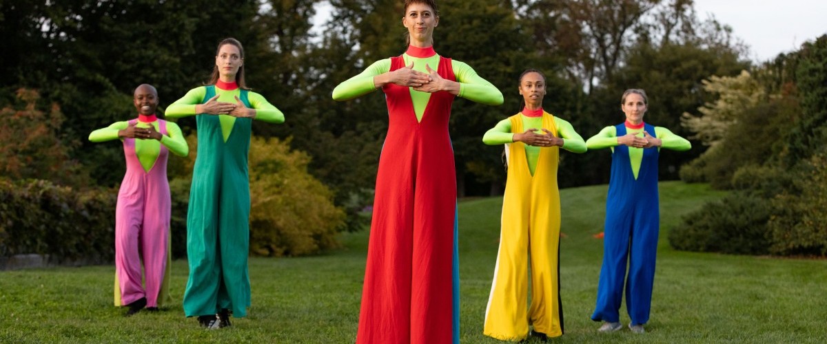 Five dancers in boldly colored pantsuits pose on a grassy lawn.