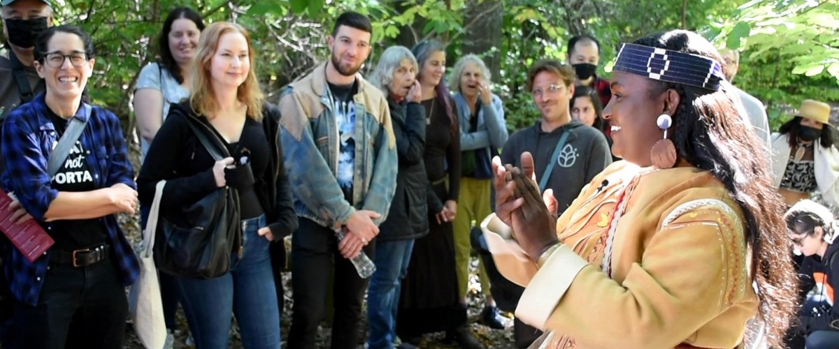 A woman in tribal dress speaks to a group of smiling people under green trees.