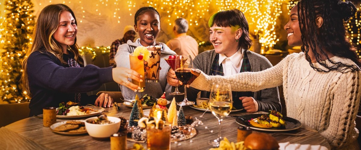 Four women toast at a festive restaurant table amid holiday lights and decor.