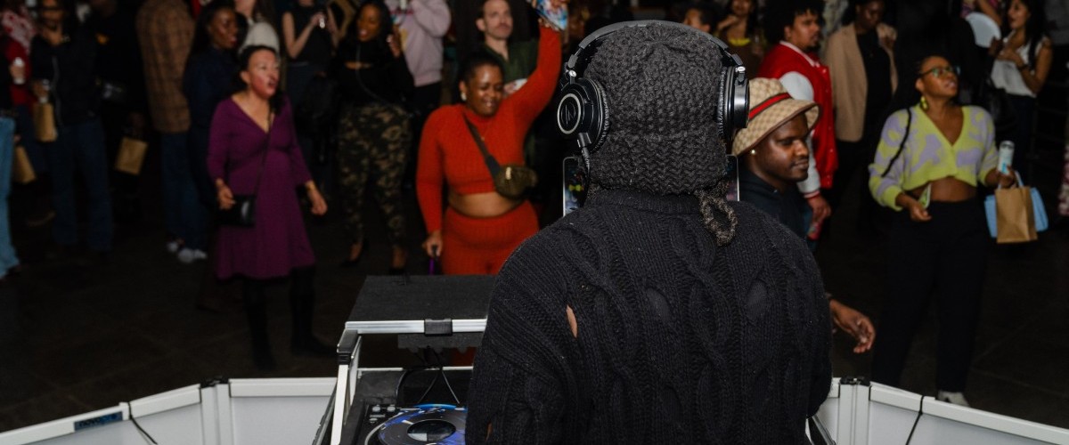 A photo from the stage behind a DJ wearing a hat and headphones in front of a dancing crowd.